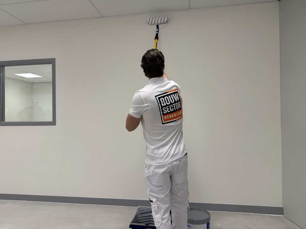 Schilder staat bij verfbak en emmer en rolt een witte wand tot vlak onder het plafond