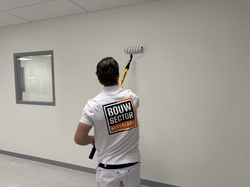 Schilder rolt een witte wand naast een binnenraam met een grijs kozijn