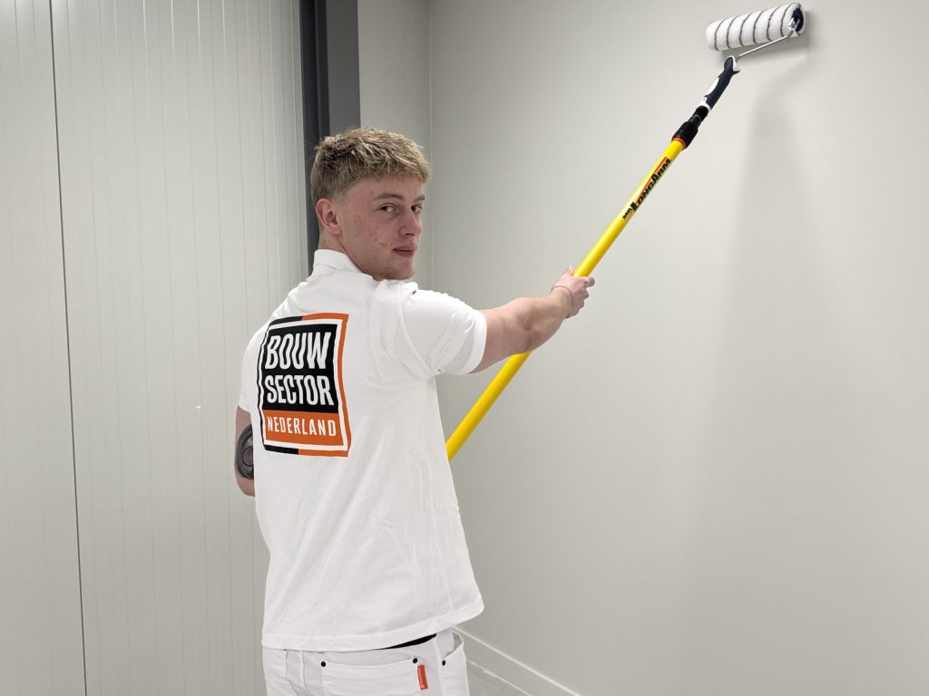 Jonge schilder kijkt over zijn schouder terwijl hij een witte muur rolt met een gele telescoopsteel