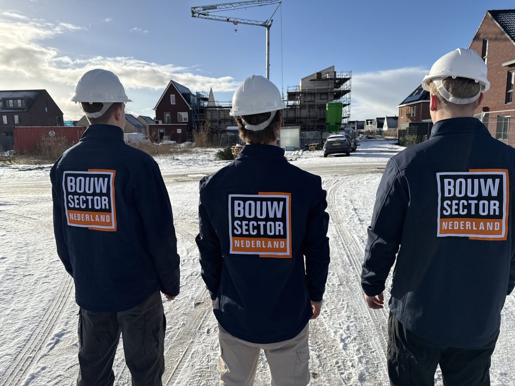 Drie vakmensen van Bouwsector Nederland met witte helm en werkjas kijken uit over een straat met woningen en steigers