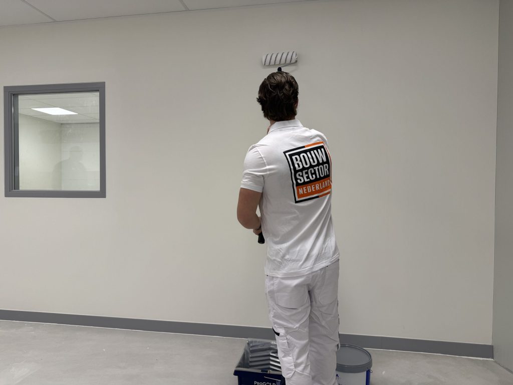 Schilder van Bouwsector Nederland rolt een lichte wand tot tegen het systeemplafond, met verfbak en emmer op de vloer