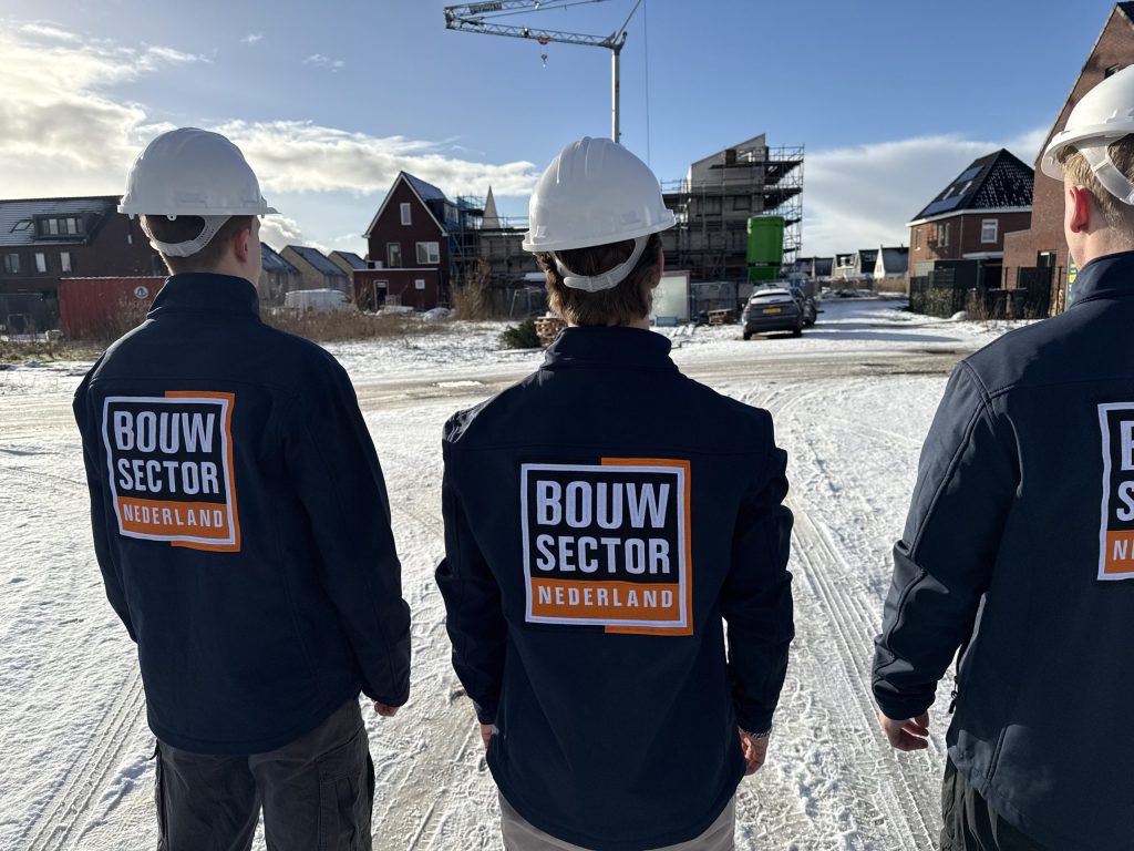 Drie vakmensen van Bouwsector Nederland met witte helm, op de rug gezien, kijken naar huizen in aanbouw in de sneeuw